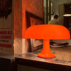 Orange Clérachic lamp lit on a rough wooden shelf in front of framed posters, its stem glowing warm.