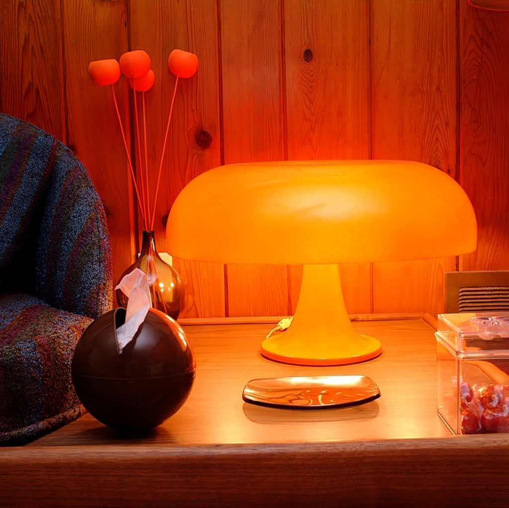 Orange Clérachic table lamp lit on a wooden side table, with orange stems in a dark vase and a plate beside it.