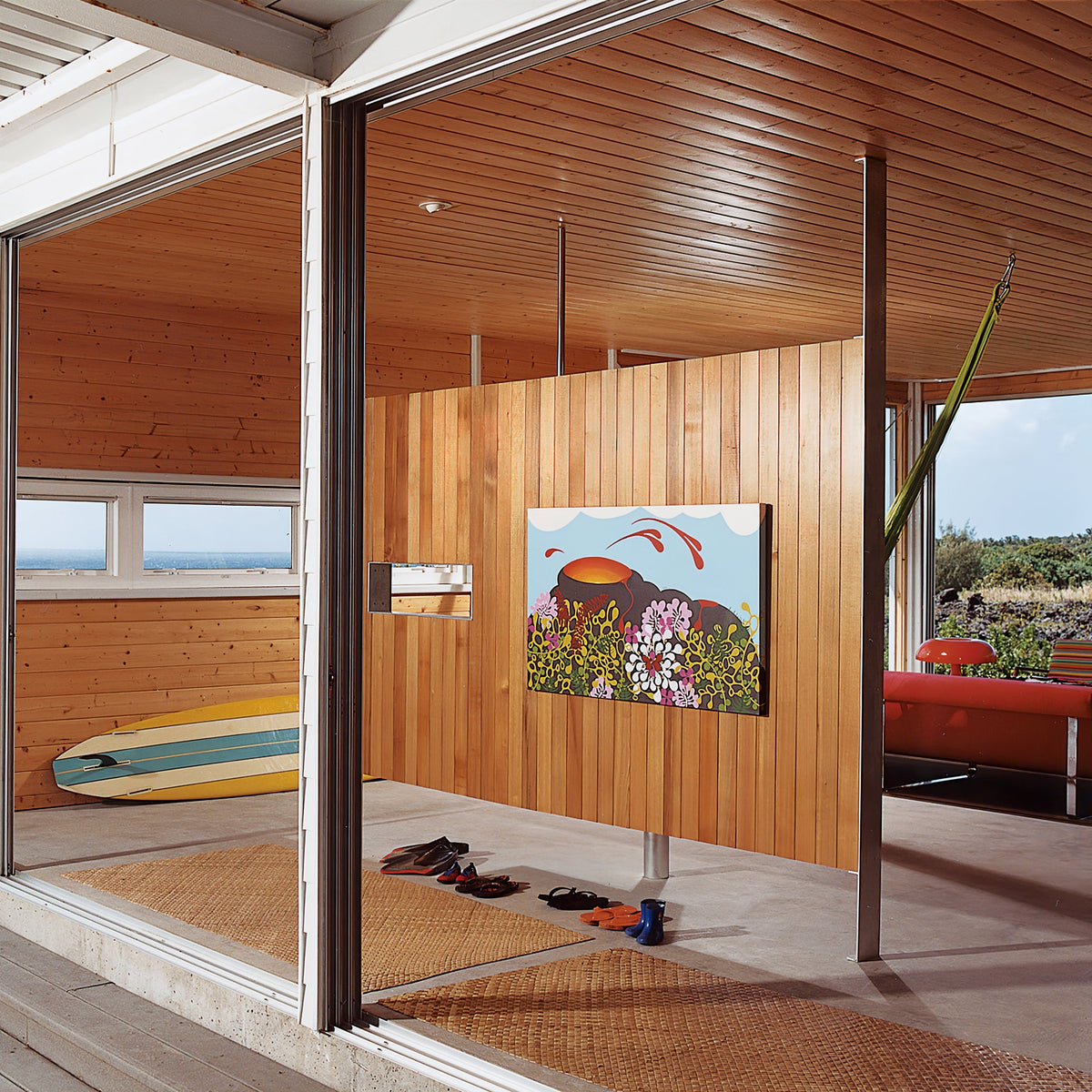 A Clérachic orange mushroom lamp seen from far across a wood-lined room with big glass doors and a surfboard on the wall.