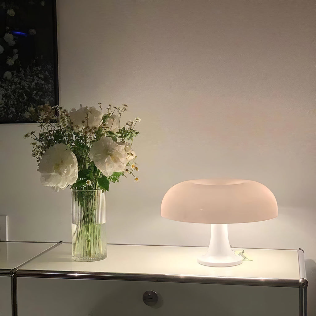 White Clérachic lamp glowing on a glass-top cabinet, with a vase of white flowers beside it against a pale wall.