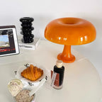 A Clérachic orange table lamp on a white table beside a black ceramic vase, a croissant and a small bottle.