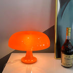 Orange Clérachic lamp lit on a white surface in front of a framed blue-green print, with a bottle of cognac beside it.