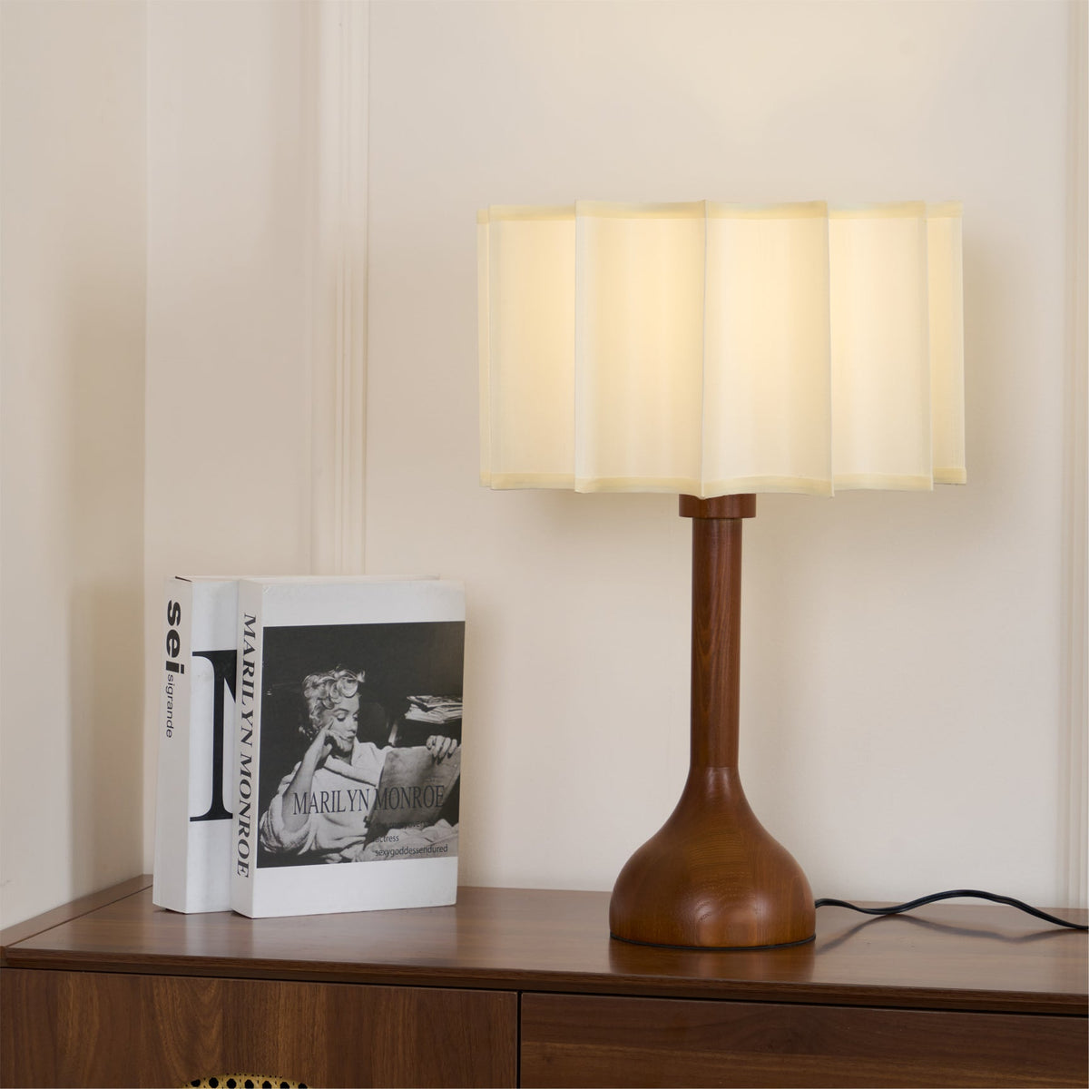 Unlit Clérachic table lamp with a wide scalloped fabric shade, standing next to a book on a wooden cabinet.