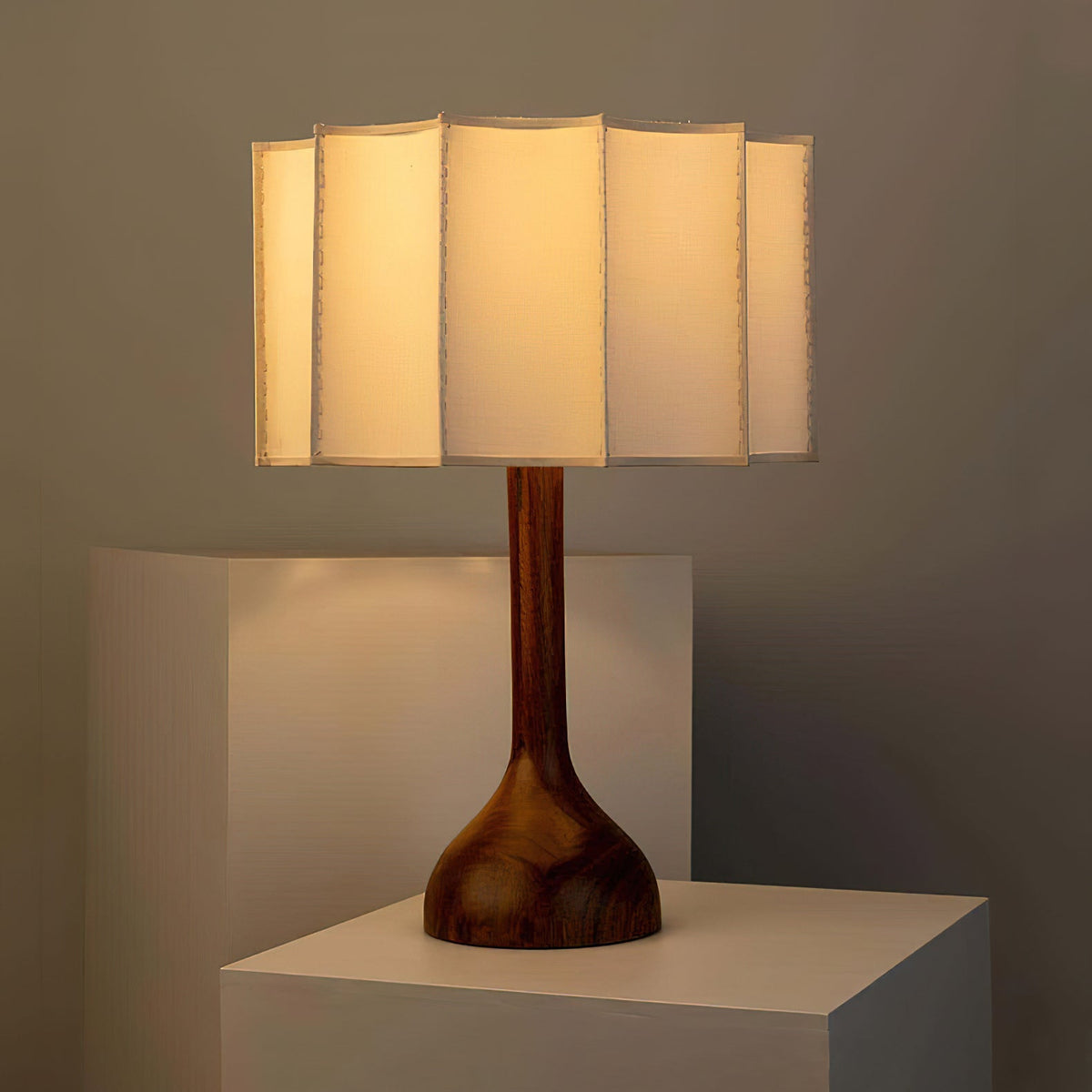 Lit Clérachic table lamp on a pale plinth against a dark grey wall, a warm glow through the flat-panelled shade.