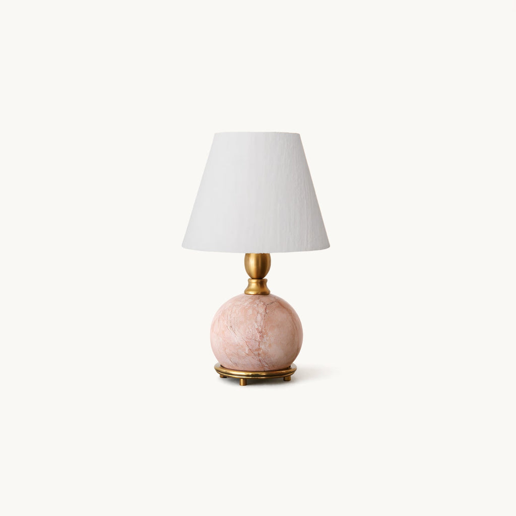 A Clérachic table lamp with a round pink marble base on a brass foot and a white linen shade, on a plain off-white backdrop.