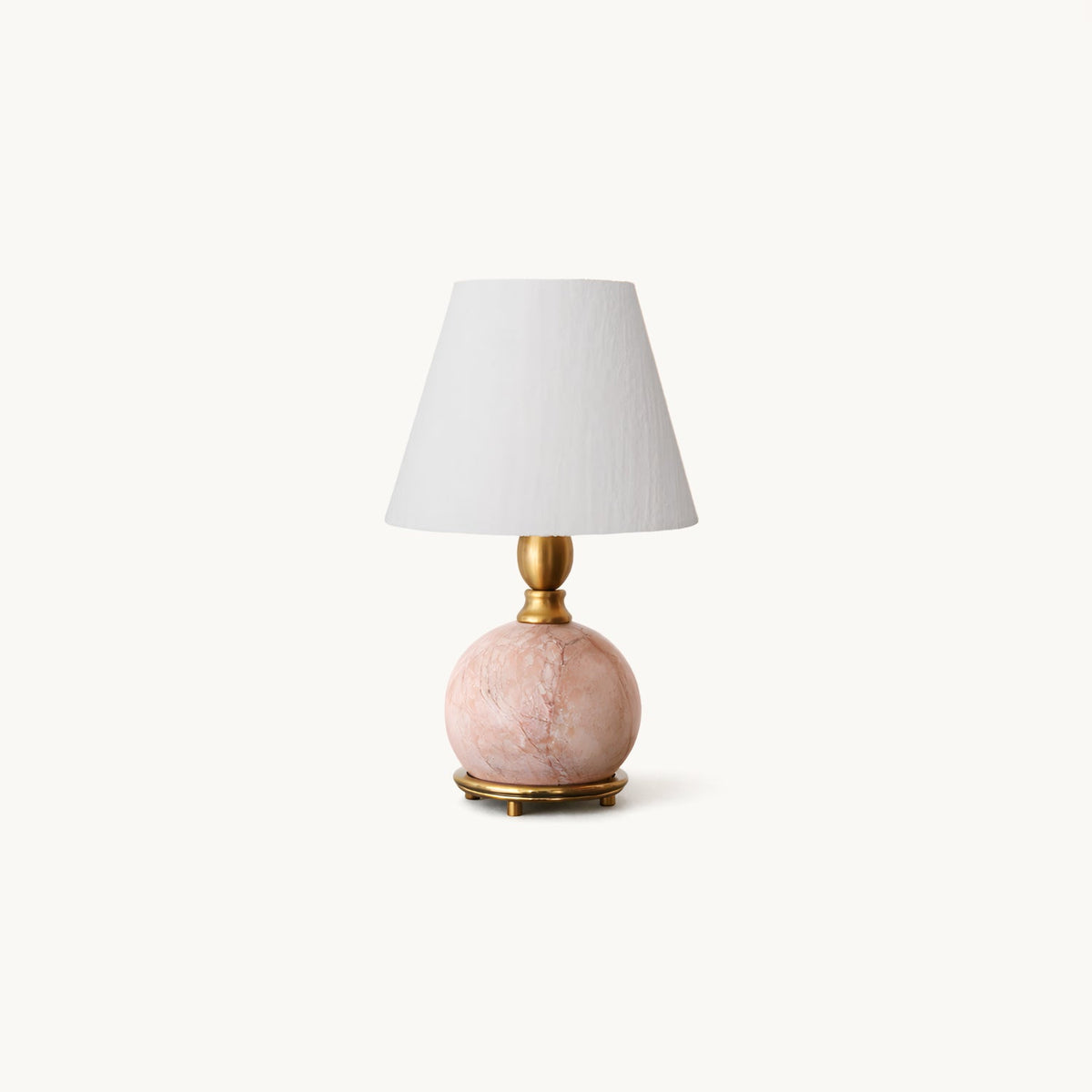A Clérachic table lamp with a round pink marble base on a brass foot and a white linen shade, on a plain off-white backdrop.