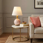 A Clérachic pink marble table lamp with a white shade stands on a round brass side table, beside a cream armchair and coral cushion.