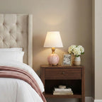 On a walnut bedside table, a Clérachic pink marble lamp with a white shade sits next to a framed photo and white flowers.