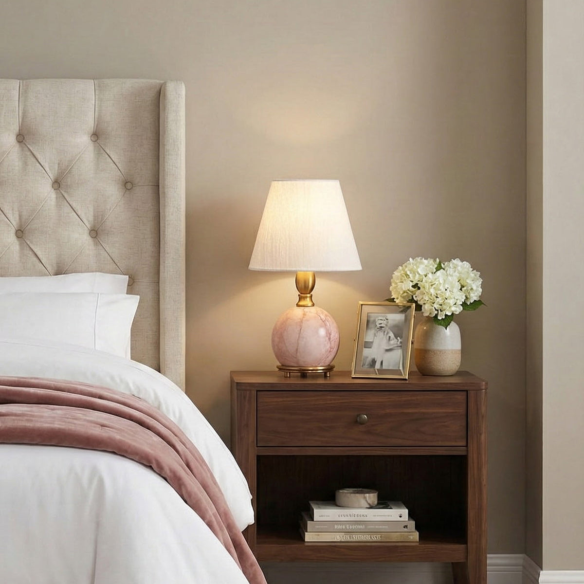 On a walnut bedside table, a Clérachic pink marble lamp with a white shade sits next to a framed photo and white flowers.