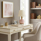 A Clérachic pink marble table lamp stands on a cream writing desk with gold trim, near a bookcase and a framed print.