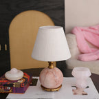 A Clérachic lamp with a round pink marble base and white shade sits on a magazine, with a pink throw blurred behind.