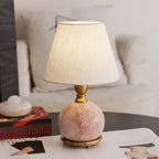 Close view of a lit white fabric shade and a round pink marble base with a brass collar, on a Clérachic table lamp.