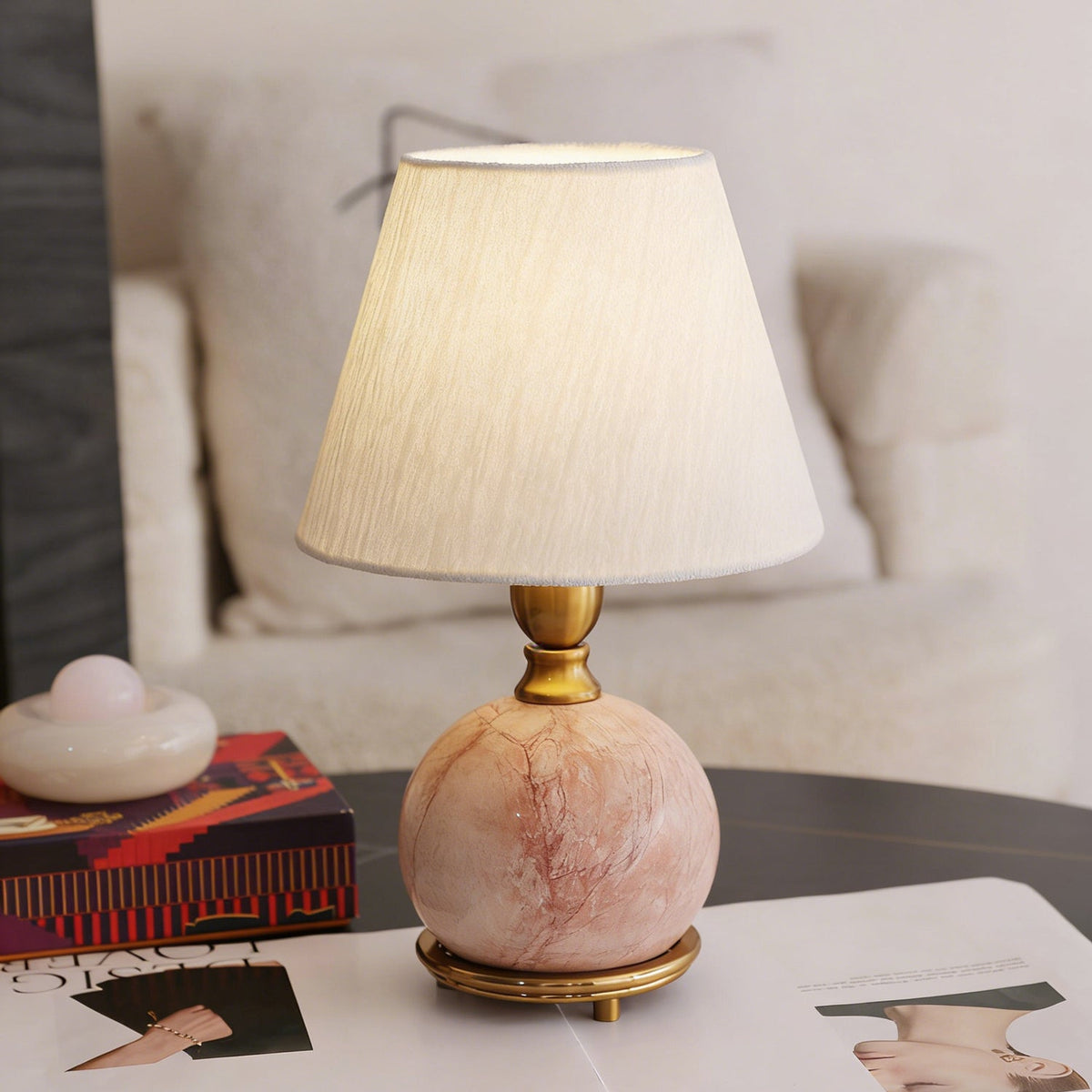 Close view of a lit white fabric shade and a round pink marble base with a brass collar, on a Clérachic table lamp.