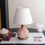 A Clérachic table lamp with a veined pink marble ball base and a crinkled white shade, on a dark table with a magazine.