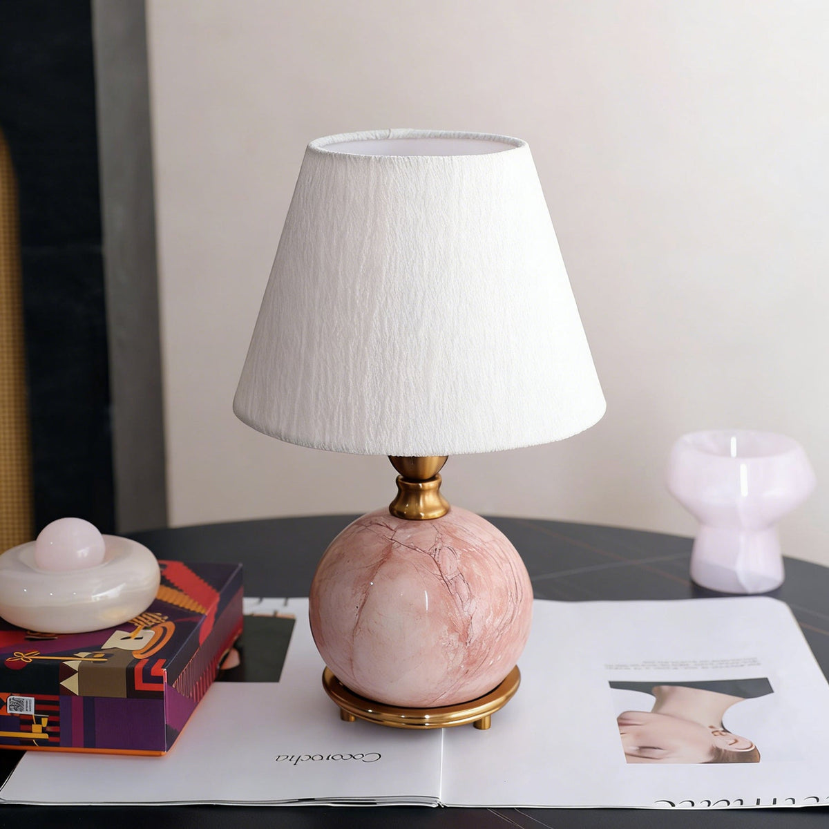A Clérachic table lamp with a veined pink marble ball base and a crinkled white shade, on a dark table with a magazine.
