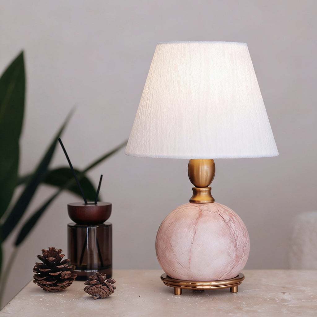 A Clérachic pink marble lamp with a lit white shade on a stone-look surface, beside a reed diffuser and two pine cones.