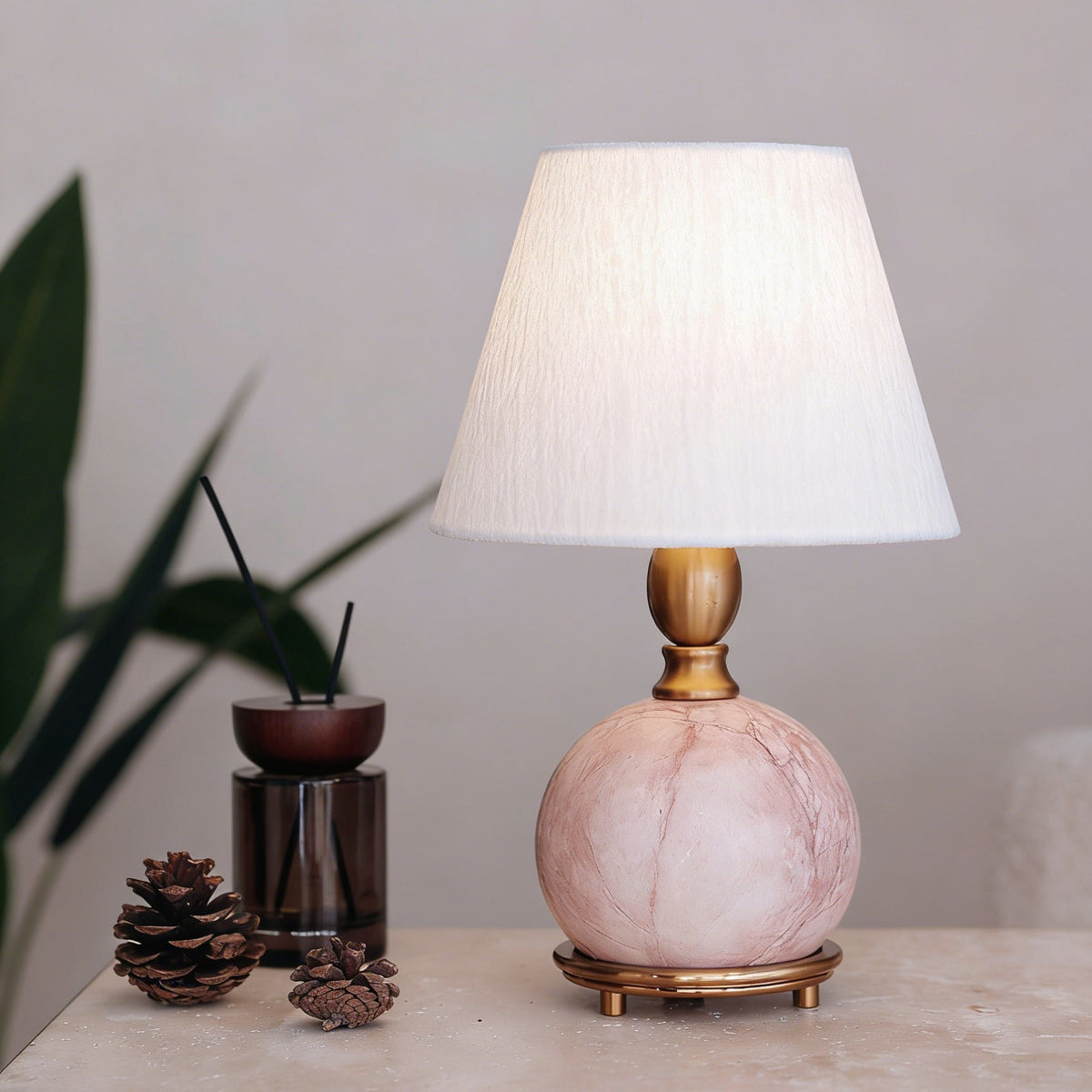 A Clérachic pink marble lamp with a lit white shade on a stone-look surface, beside a reed diffuser and two pine cones.