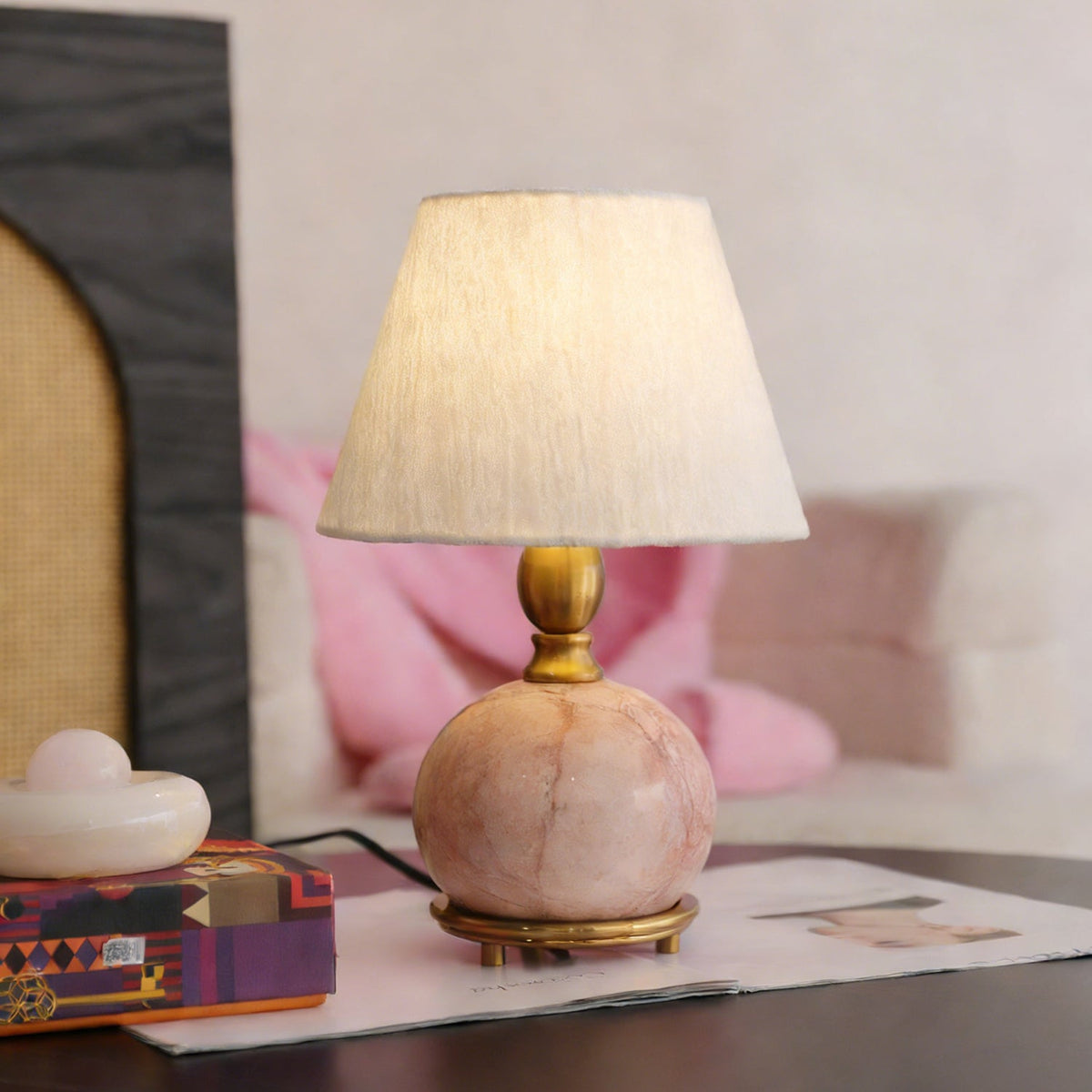 A lit Clérachic table lamp with a pink marble base, on a dark table in front of a rattan arch and a pink throw.