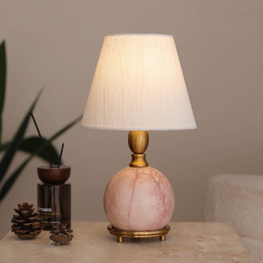 A Clérachic table lamp with a glowing white shade and pink marble base, beside a diffuser, pine cones and a dark plant.