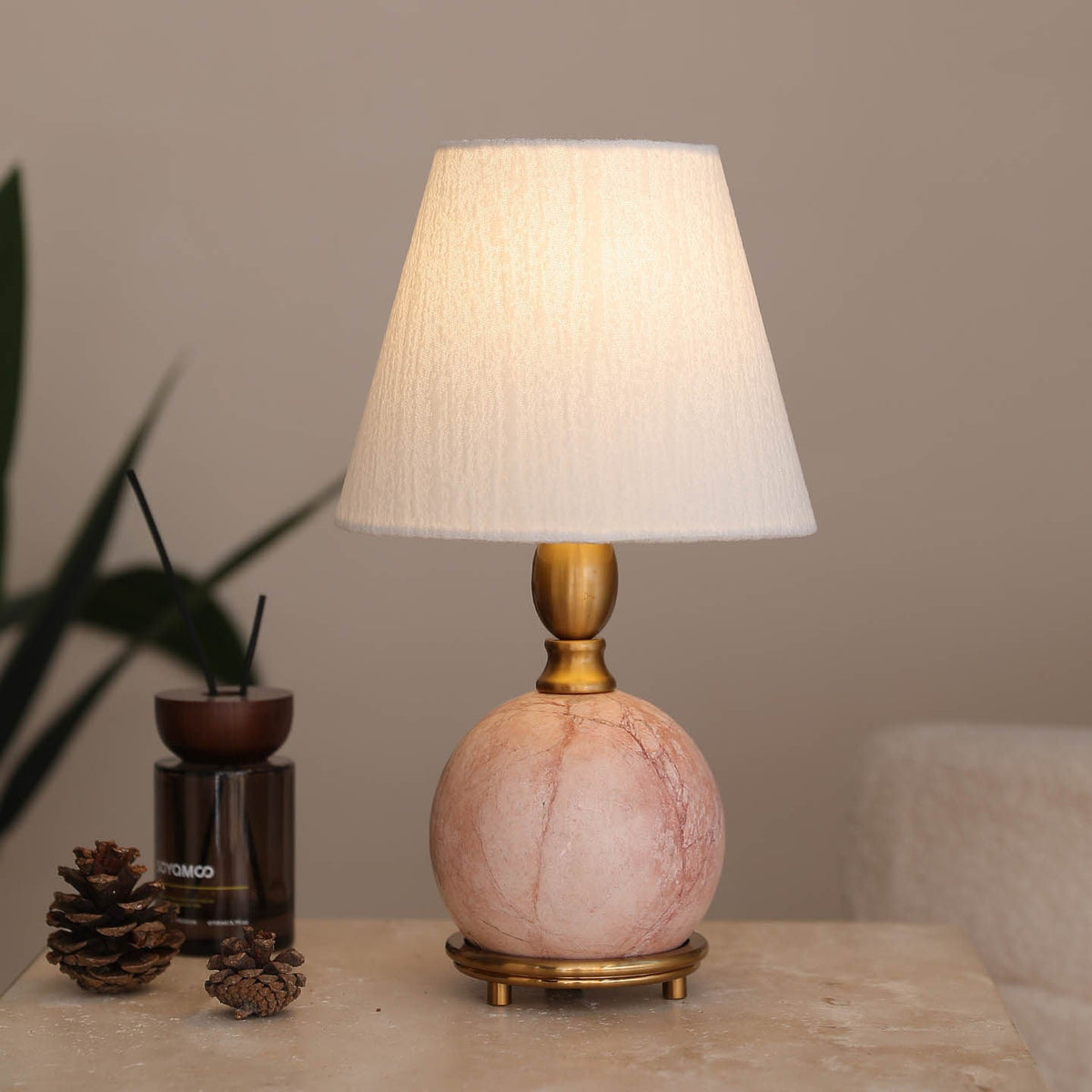A Clérachic table lamp with a glowing white shade and pink marble base, beside a diffuser, pine cones and a dark plant.