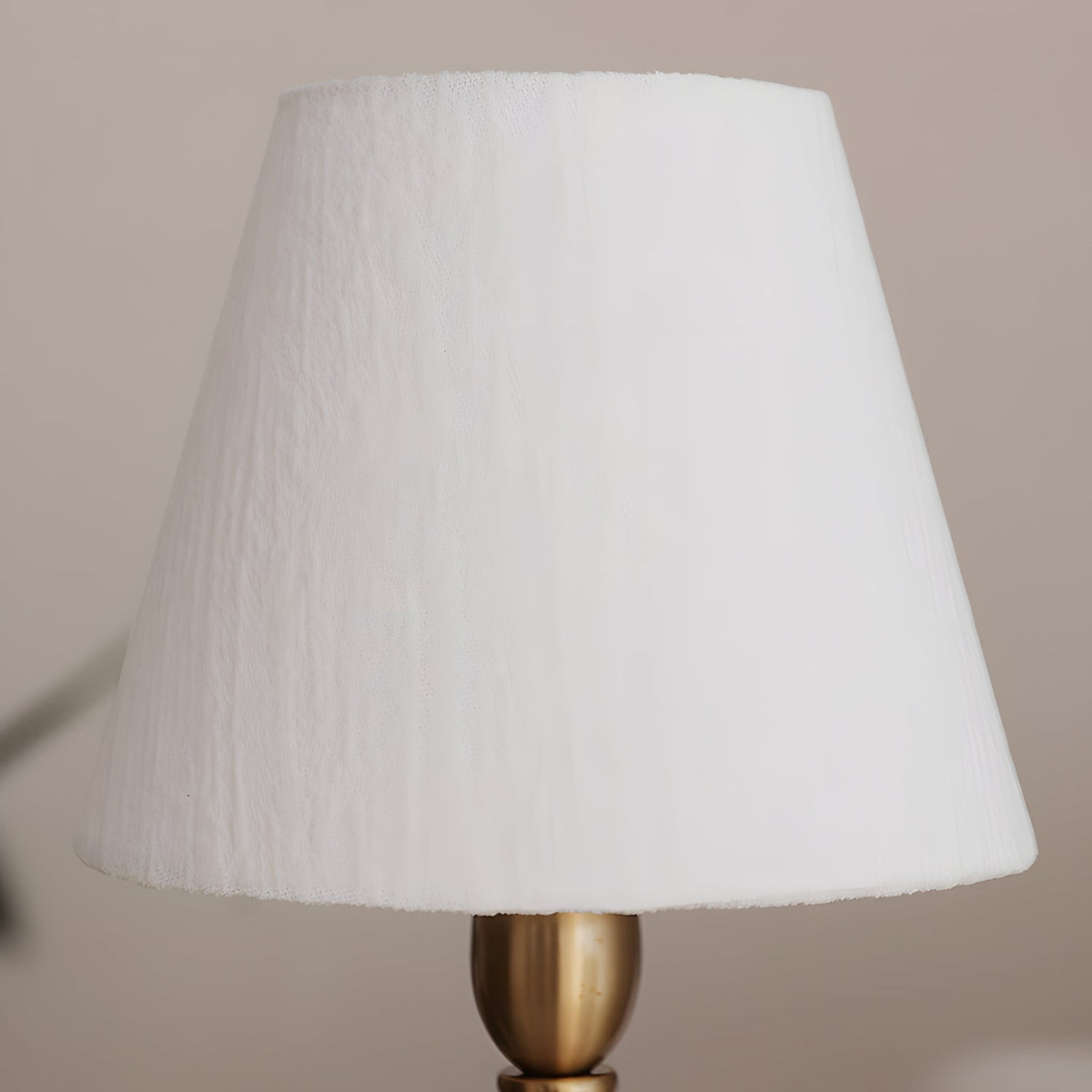 Close view of the crinkled white fabric shade on this Clérachic lamp, above its brass neck and a mauve-grey wall.