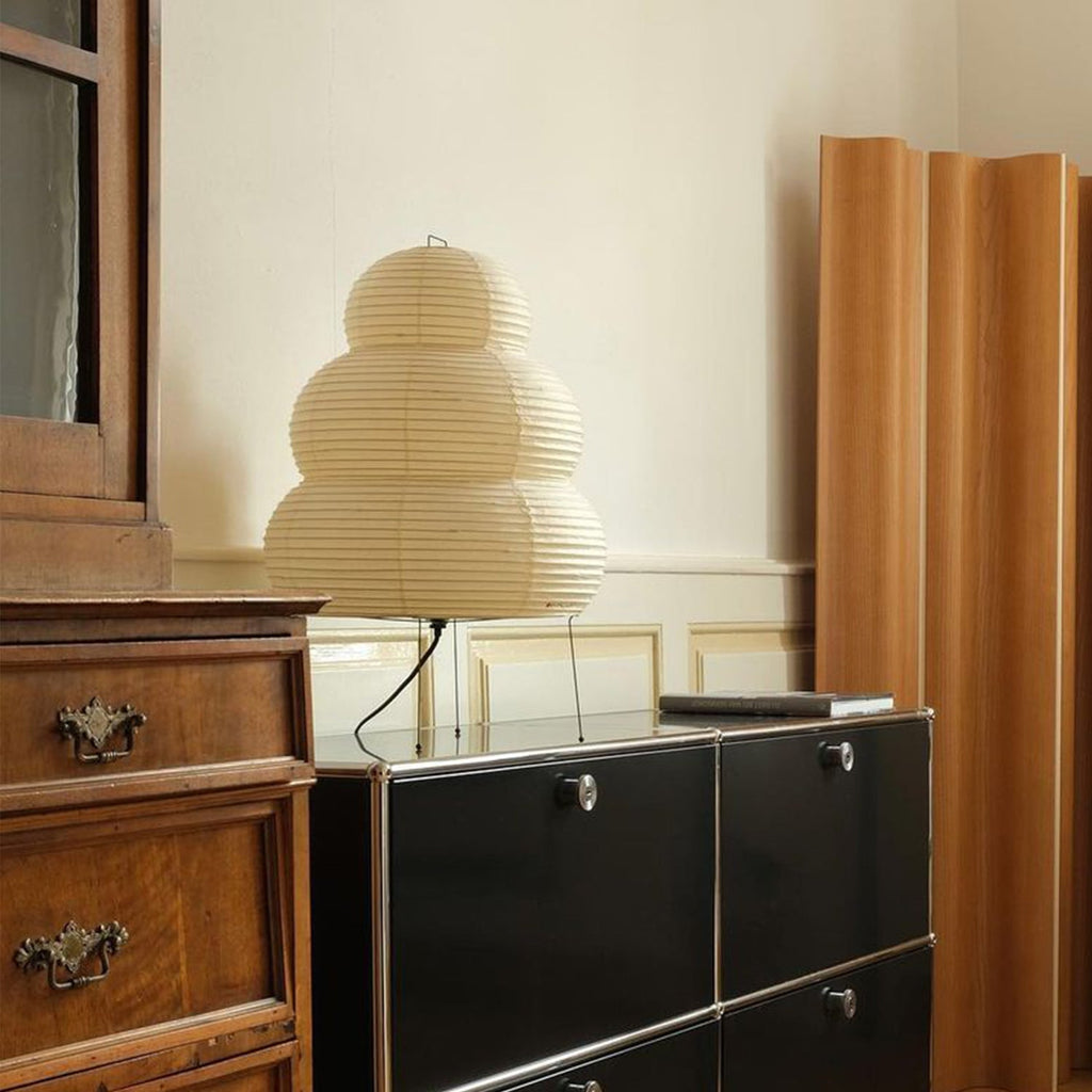 Tiered paper shade of this Clérachic table lamp, unlit, on a black cabinet beside a pale wooden room divider.