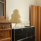 Tiered paper shade of this Clérachic table lamp, unlit, on a black cabinet beside a pale wooden room divider.