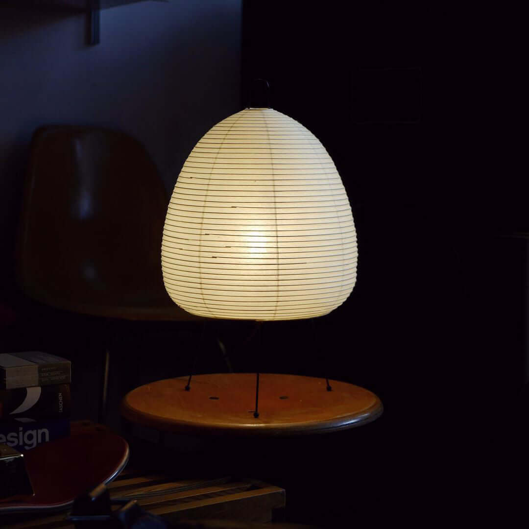 In a dark room, this Clérachic paper lamp glows as the only light, sitting on a round wooden stool.