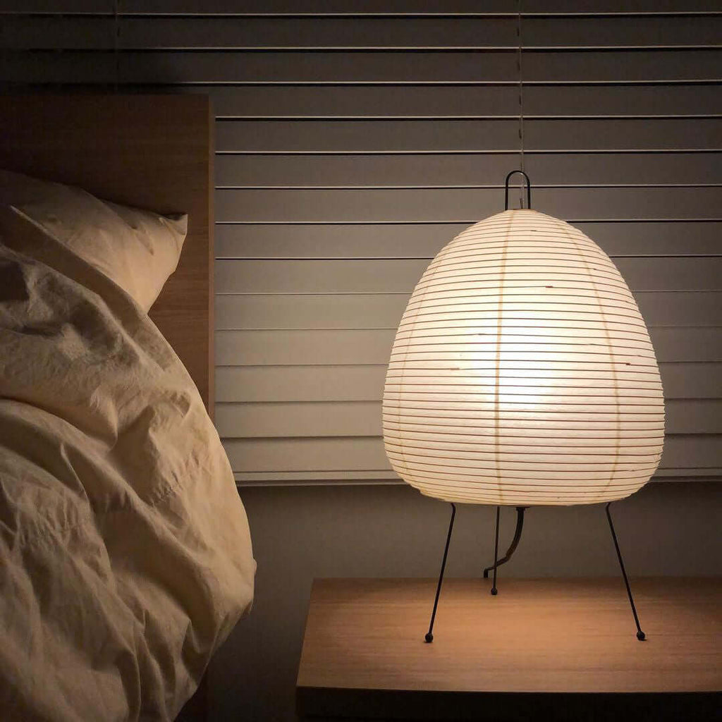 A Clérachic egg-shaped paper lamp lit on a bedside table, with rumpled linen at the left and window blinds behind.