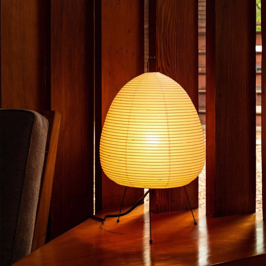 Warm amber glow from this Clérachic paper lamp on a polished wooden floor, beside an armchair and slatted wood panelling.