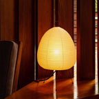 Warm amber glow from this Clérachic paper lamp on a polished wooden floor, beside an armchair and slatted wood panelling.