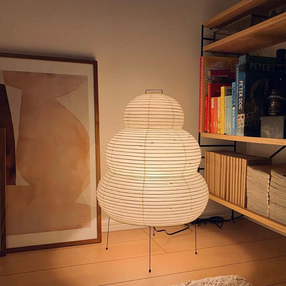 A tall Clérachic tiered paper lamp lit on the floor between a framed abstract print and a shelf of books.