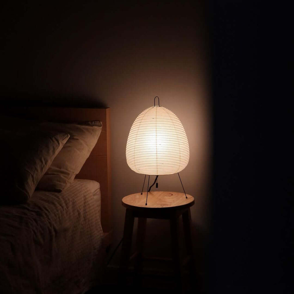 Small Clérachic paper lamp lit on a round wooden stool beside a bed in a dark bedroom, giving the only light.