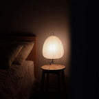 Small Clérachic paper lamp lit on a round wooden stool beside a bed in a dark bedroom, giving the only light.