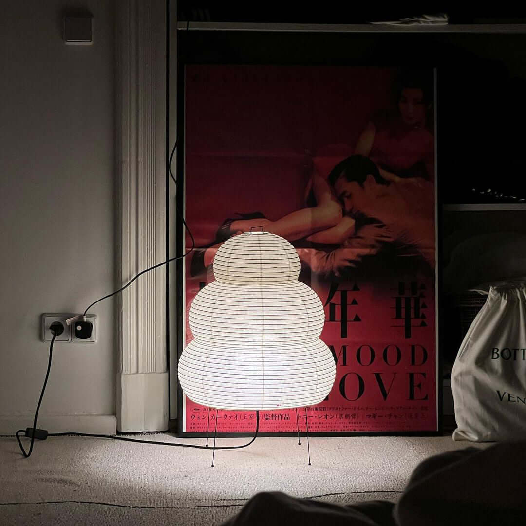 This Clérachic tiered paper lamp glows on the floor in front of a red film poster, its cord running to a wall socket.