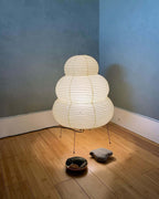 A lit Clérachic tiered paper lamp in the corner of a room with a blue wall, standing on a bamboo floor beside two stones.
