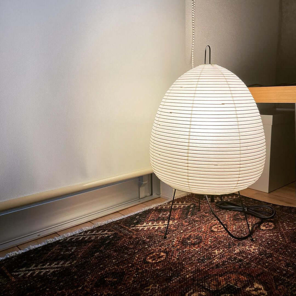 Large Clérachic egg-shaped paper lamp on a patterned rug against a pale wall, its black cord curling across the floor.