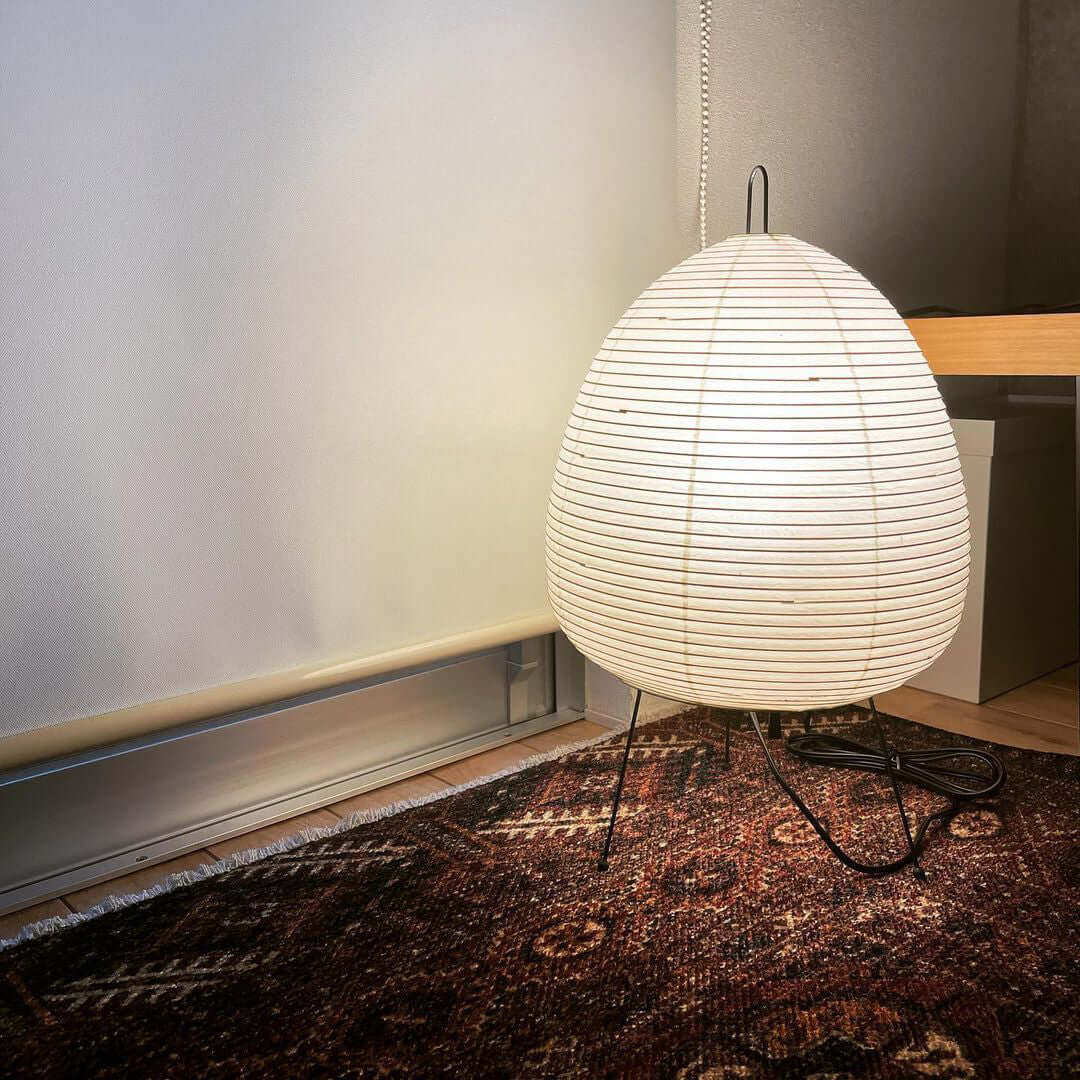 Large Clérachic egg-shaped paper lamp on a patterned rug against a pale wall, its black cord curling across the floor.