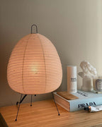 This Clérachic paper lamp shows a rosy glow on a pale wooden shelf, beside a stack of books and a candle.