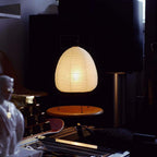 Dimly lit corner where this Clérachic paper lamp glows on a round wooden stool, with shelves and speakers behind it.