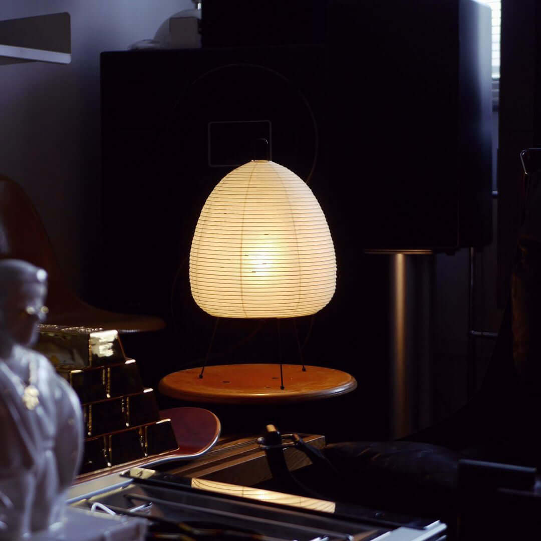 Dimly lit corner where this Clérachic paper lamp glows on a round wooden stool, with shelves and speakers behind it.