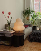 Tiered Clérachic paper lamp lit on a black side table in a living room, next to a sofa, houseplants and a vase of tulips.