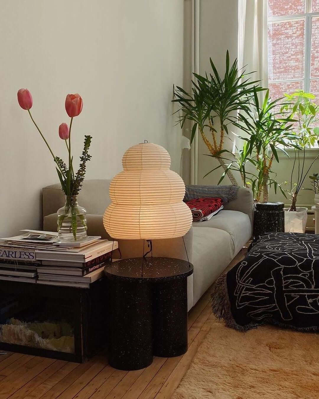 Tiered Clérachic paper lamp lit on a black side table in a living room, next to a sofa, houseplants and a vase of tulips.
