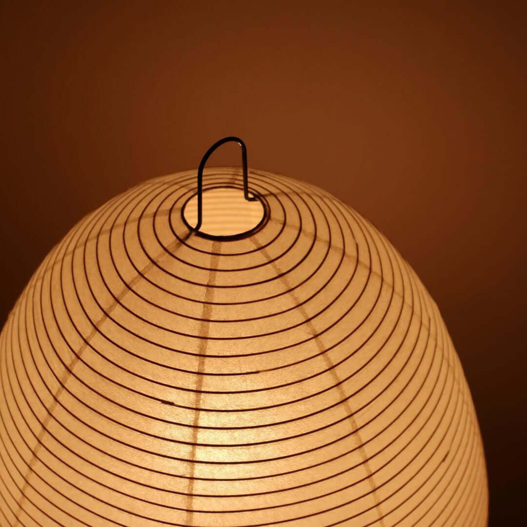 Close view of the ribbed paper shade and black wire hanging loop on this Clérachic lamp, glowing amber.