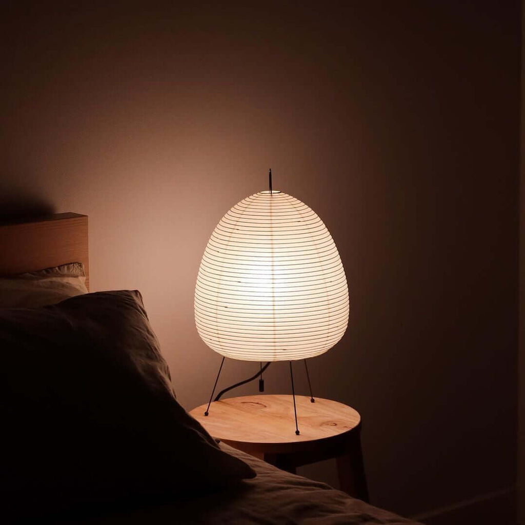 A Clérachic egg-shaped paper lamp glows on a small wooden stool beside a bed, in a dim and quiet room.