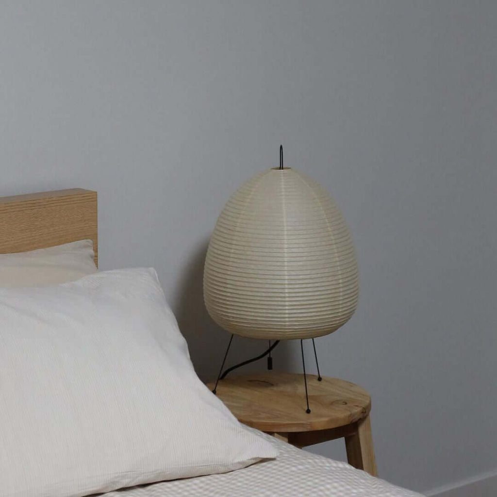 This Clérachic paper lamp, unlit, sits on a round wooden stool next to a bed dressed in white linen.