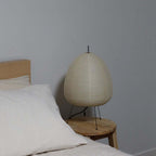 This Clérachic paper lamp, unlit, sits on a round wooden stool next to a bed dressed in white linen.