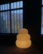 A tiered Clérachic paper lamp glows amber on the floor in front of a tall steel-framed window at dusk.