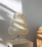 Sunlight casts shadows across a tiered Clérachic paper lamp, switched off, on a white block against a grey wall.