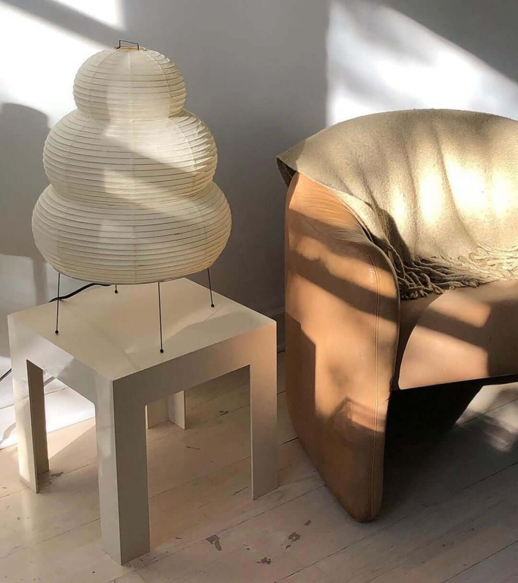 This Clérachic tiered paper lamp stands on a white side table next to a tan leather armchair, lit by afternoon sun.